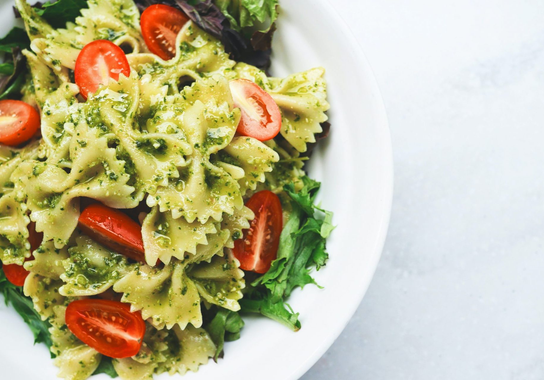 Farfalle with pesto, green salad and tomatoes in a white bowl on a light background