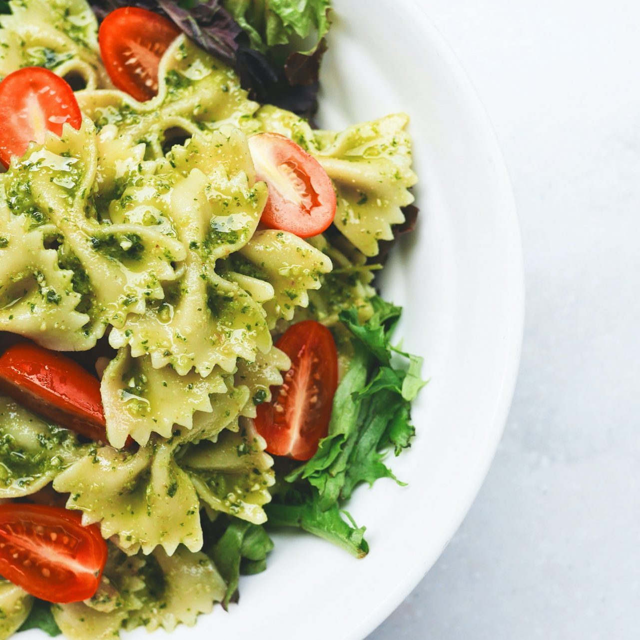 Farfalle with pesto, green salad and tomatoes in a white bowl on a light background