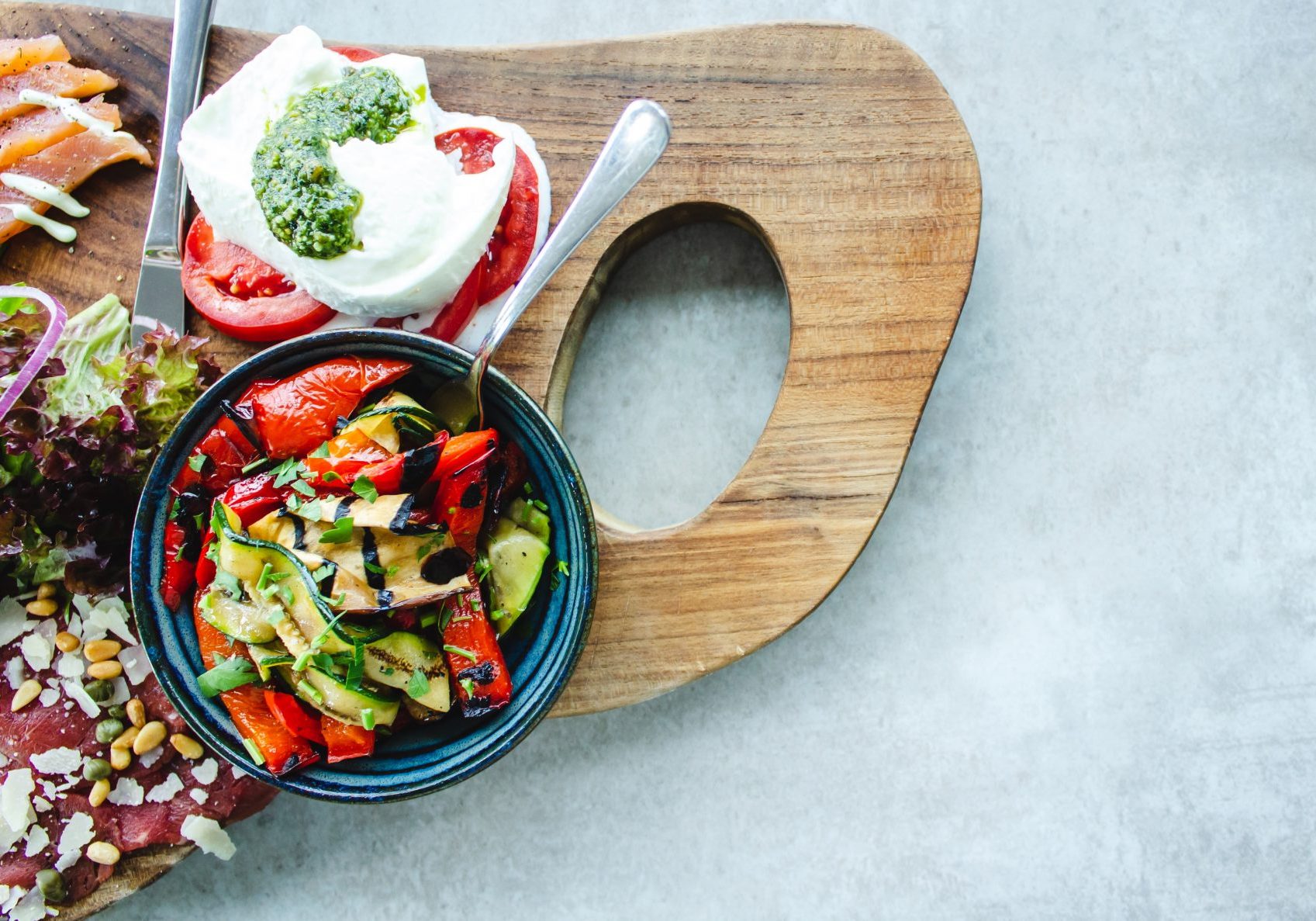 Small bowl with grilled vegetables and a dressing on top. Next to it mozzarella and tomatoes with pesto on a wooden plate on a light background