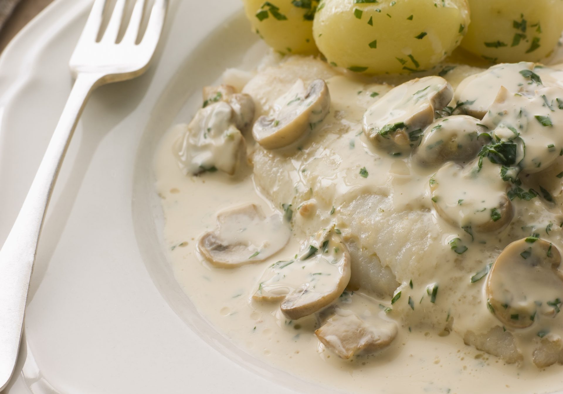 Fish fillet with potatoes and mushroom sauce on a white plate, fork on the side and napkin on wooden table